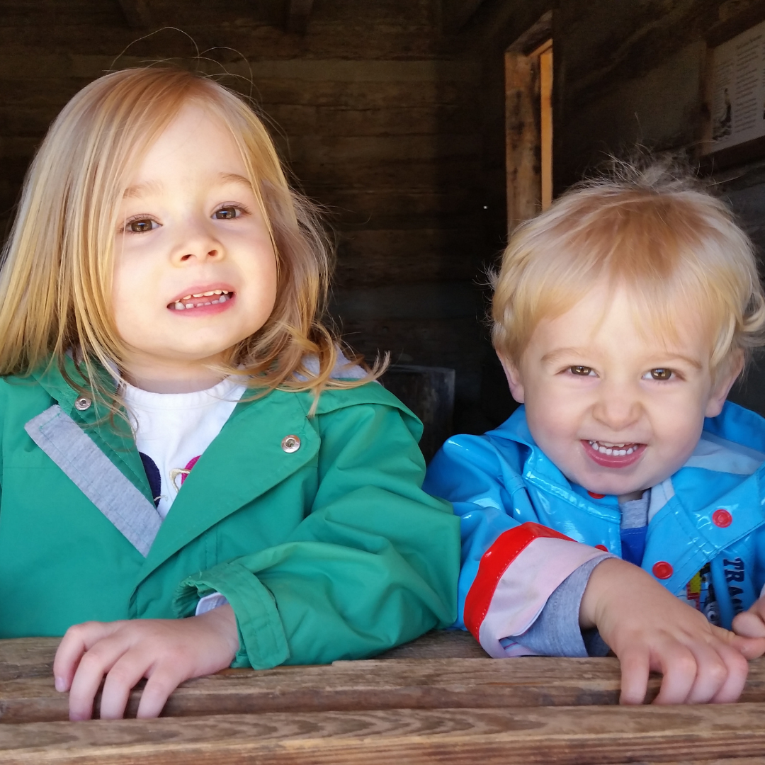 A young blonde girl and a younger blonde  boy peek out from a wooden structure. The girl is wearing a green coat and the boy is wearing a blue coat. 